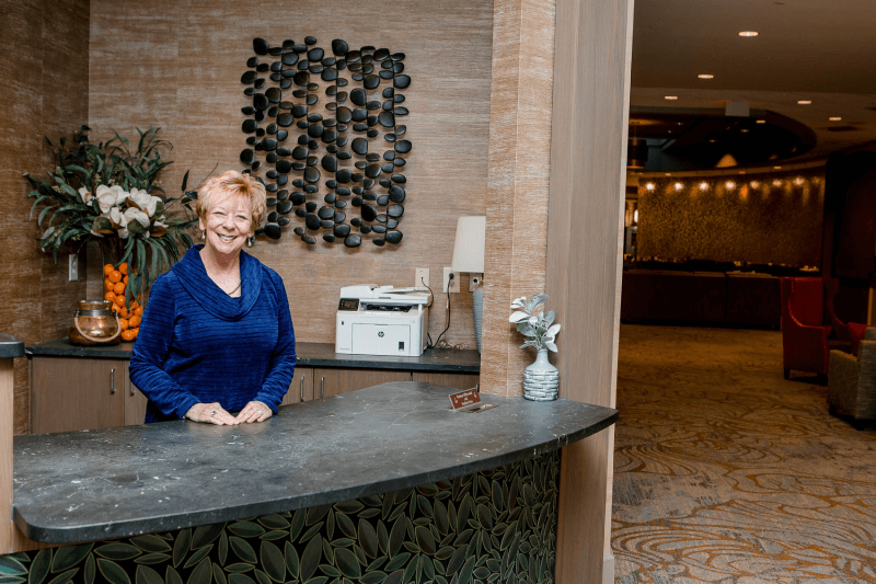 A woman stands behind a reception desk in a hotel lobby, smiling at the camera. There is a decorative wall piece and a printer behind her, and the lobby is visible in the background.