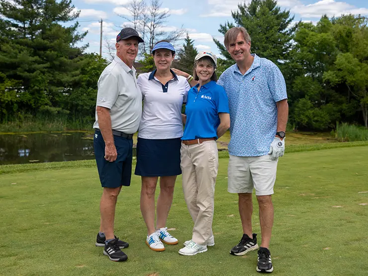 Four adults stand together on a golf course, dressed in golf attire, posing for a group photo with trees and blue sky in the background.