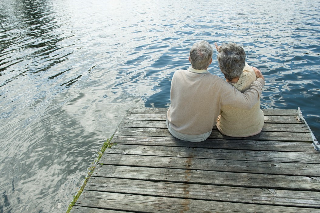 Two elderly people sit close together on a wooden dock, facing a large body of water. One has an arm around the other, who is pointing out over the lake.