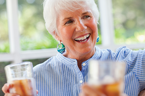 Smiling older woman with short white hair and blue shirt holds a glass of iced tea, sitting indoors by a window, with another glass in the foreground.