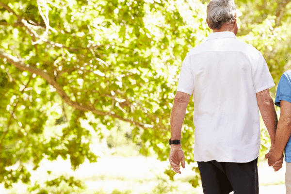 An older couple holding hands walks outdoors on a sunny day, surrounded by green trees and bright natural light.