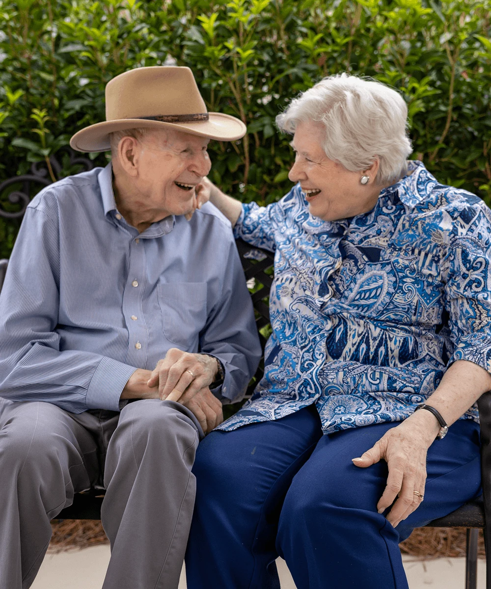 Two elderly people sit on a bench outdoors, smiling and holding hands. Both wear blue clothing and appear to be enjoying a friendly conversation. Green bushes are visible in the background.