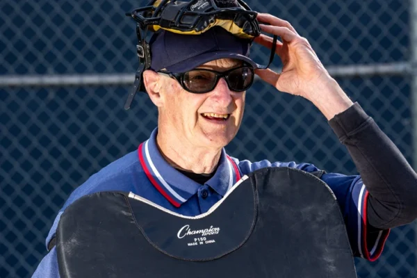 Older baseball umpire wearing sunglasses, a face mask on top of his cap, and protective gear, smiling in front of a chain-link fence.