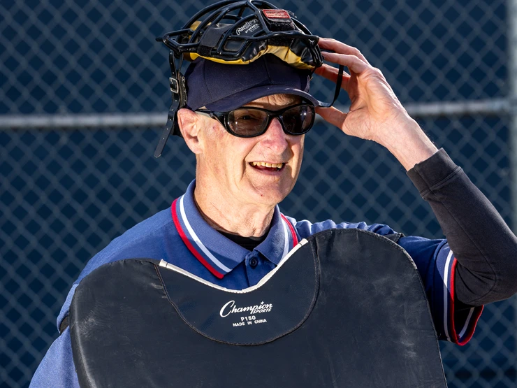 Older baseball umpire wearing sunglasses, a face mask on top of his cap, and protective gear, smiling in front of a chain-link fence.