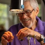 An older man with glasses and a purple shirt smiles while tying a fishing fly under a lamp, using a vise and tools.