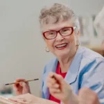 An older woman with gray hair and red glasses smiles while holding a paintbrush, sitting at a table in a well-lit indoor setting.