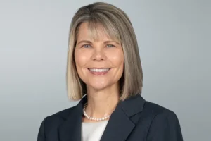 Woman with straight, shoulder-length blonde hair wearing a dark blazer, white top, and pearl necklace, smiling at the camera against a plain light gray background.