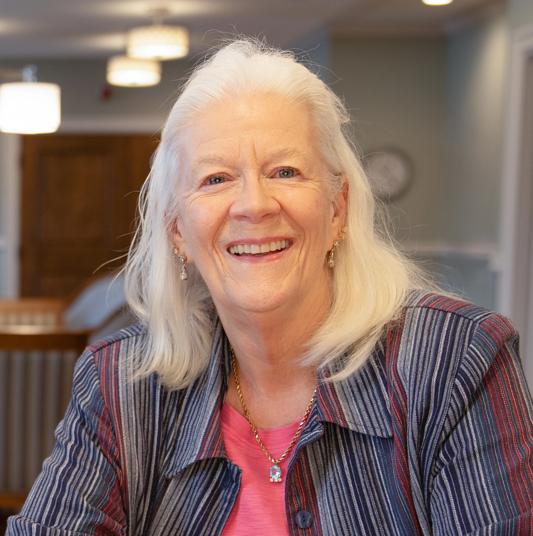 An older woman with long white hair smiles at the camera while seated indoors, wearing a striped jacket over a pink top.