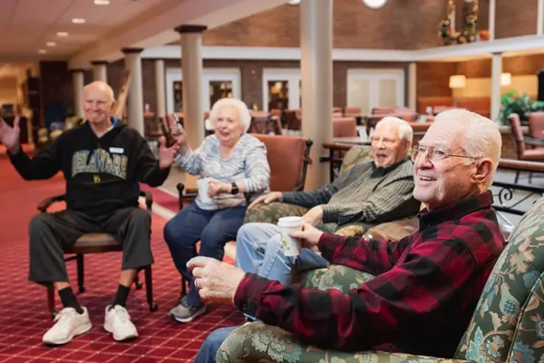 Four older adults sit and smile together in a spacious, carpeted lounge area with armchairs and coffee cups, enjoying a social gathering.