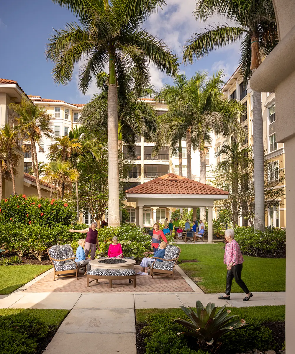 A group of seniors sit and talk on outdoor sofas under palm trees in a landscaped courtyard with a gazebo and tall residential buildings in the background.