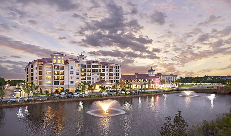 A large, multi-story building with lit windows stands by a pond with two fountains, under a cloudy evening sky.