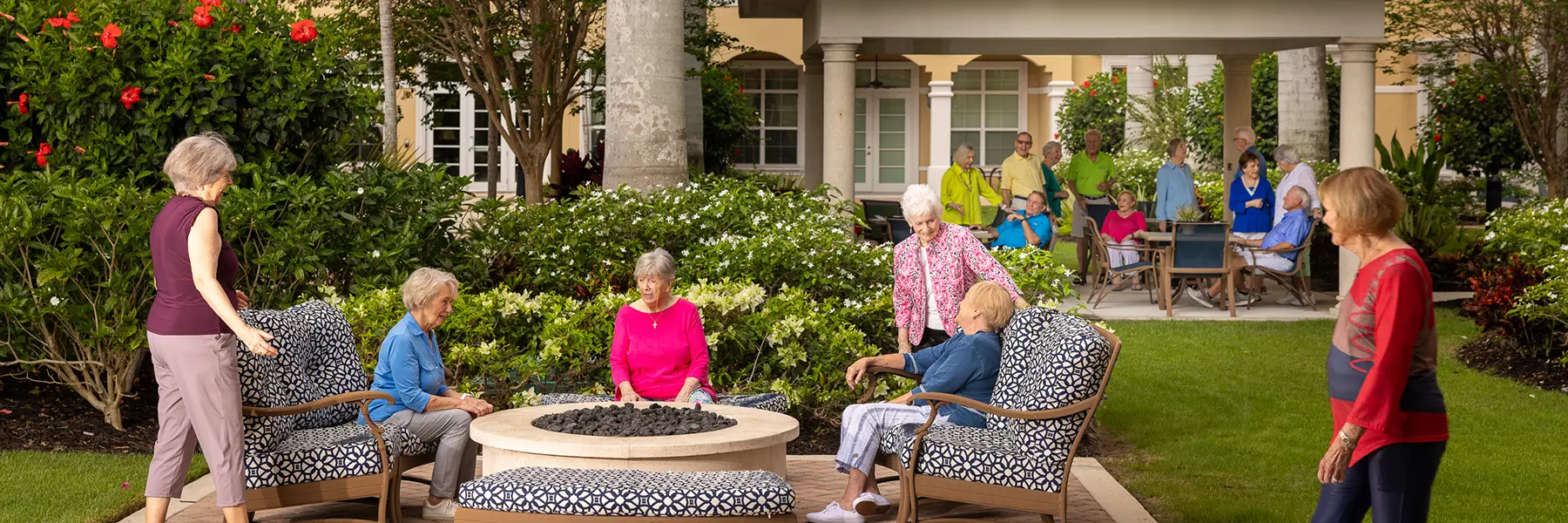 A group of older adults sit and stand around a fire pit and patio area in a landscaped outdoor setting near a building.