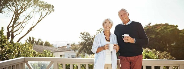 An older couple stands on a sunny balcony, smiling and holding mugs, with trees and rooftops visible in the background.