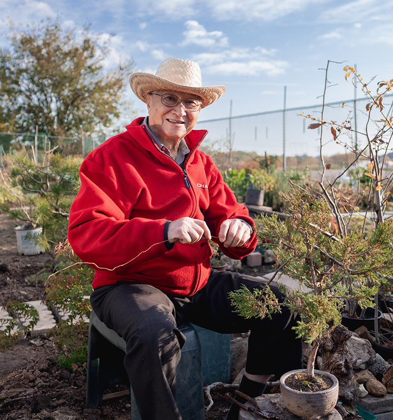 An elderly person in a straw hat and red jacket sits outdoors, smiling while pruning a small potted tree in a garden.