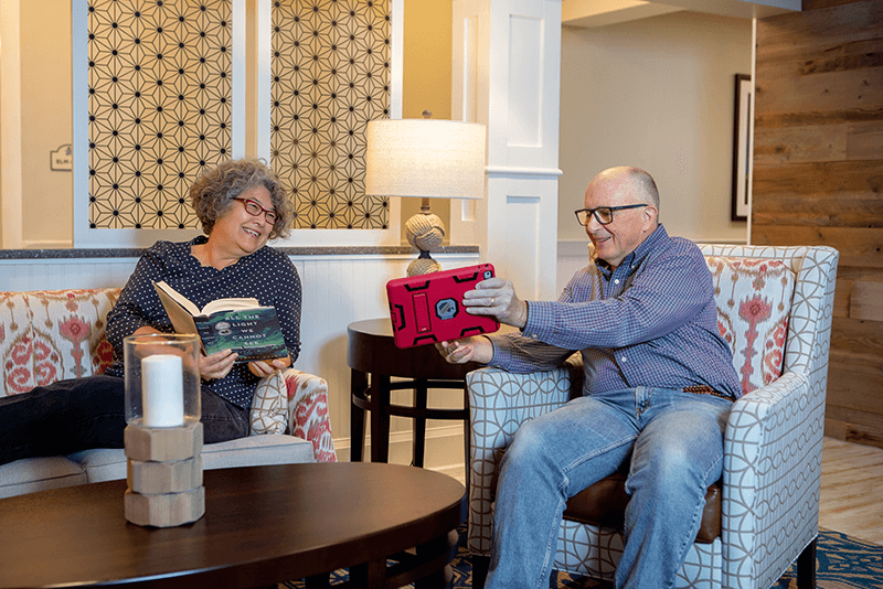 An older woman sits reading a book while a man beside her smiles and holds up a tablet; both are seated in patterned armchairs in a cozy living room.