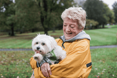 An older adult wearing a yellow jacket smiles while holding a small white dog outside in a green park.