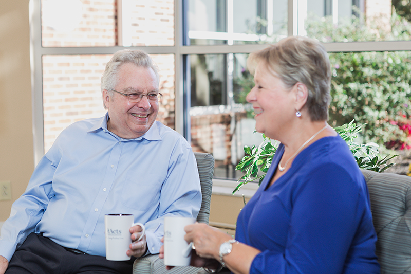 Two older adults sit indoors, smiling and talking while holding coffee mugs. Large windows and green plants are visible in the background.