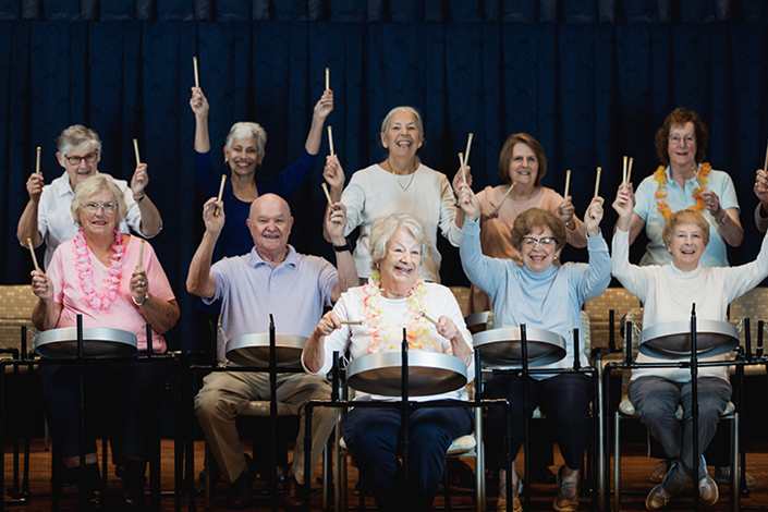 A group of older adults sits in chairs, holding drumsticks above hand drums, smiling at the camera on a stage with a blue curtain backdrop.