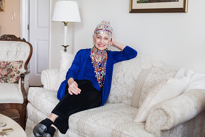 An older woman wearing a colorful crown and multiple necklaces sits on a patterned couch in a living room, smiling at the camera.