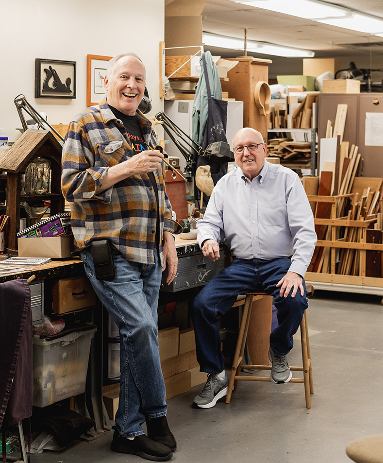 Two older men pose in a woodworking or craft studio filled with tools, wood, and supplies. One stands by a workbench, smiling, while the other sits on a stool.