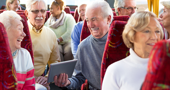 A group of older adults sit together on a bus, talking and smiling. One person holds a tablet. The seats are red with patterns.