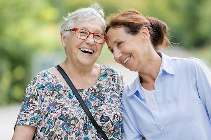 Two women, one older with grey hair and glasses, the other younger with brown hair, smile and lean close together outdoors.