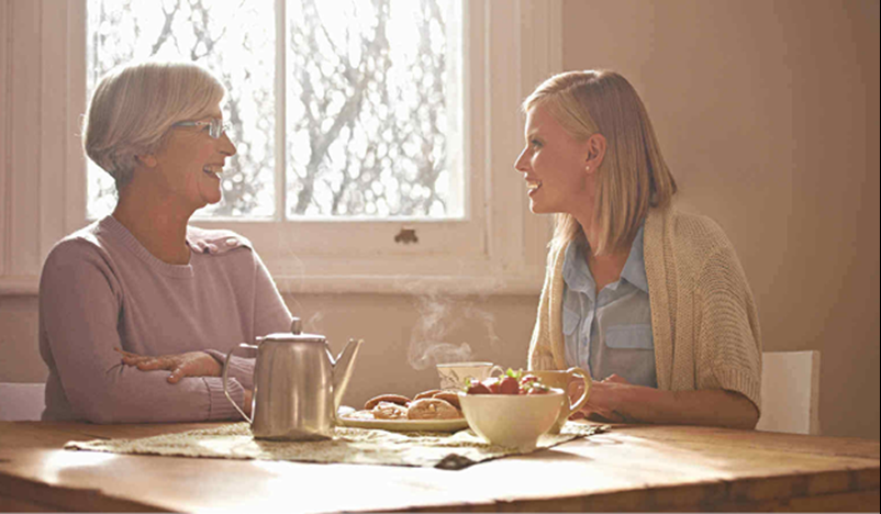 Two women sit at a table with tea, pastries, and fruit, having a conversation in a sunlit room with a window in the background.