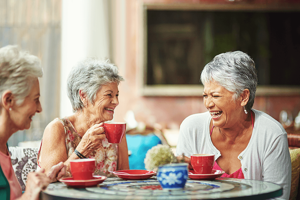 Three older women sit at a round table, smiling and laughing while drinking from red mugs outdoors.
