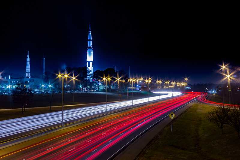Night view of a highway with red and white light trails from vehicles, streetlights, and a large illuminated rocket displayed upright in the background.