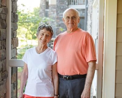An older woman and man stand side by side on a porch, smiling at the camera. Sunlight and greenery are visible in the background.
