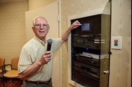 An older man holds a microphone and adjusts a control panel in a wall-mounted equipment rack in a small room.