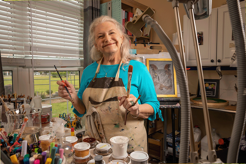 An older woman smiles while standing in an art studio, holding paintbrushes and wearing a paint-splattered apron. Jars of paint and painting supplies are visible on the table in front of her.