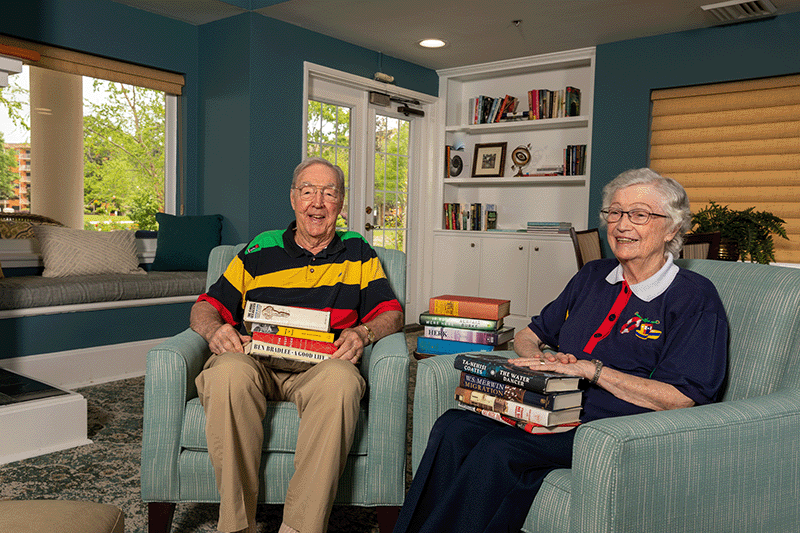 Two older adults sit in armchairs, each holding stacks of books, in a cozy living room with bookshelves, windows, and teal walls.