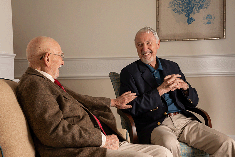Two older men sit side by side indoors, smiling and conversing. One wears a brown suit and glasses; the other wears a navy blazer and beige pants. Art decor hangs on the wall behind them.