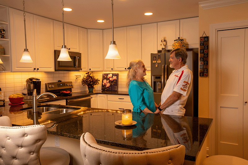 An older couple stands facing each other and smiling in a modern kitchen with white cabinets, granite countertops, and warm lighting.