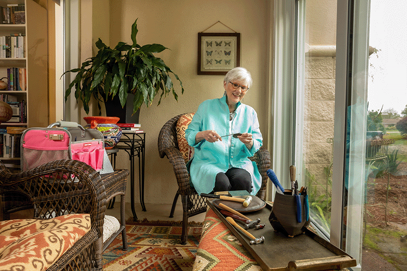 An older woman sits in a sunroom on a wicker chair, smiling while handling jewelry tools. A tray of tools is on a table nearby, with a garden visible through the window.