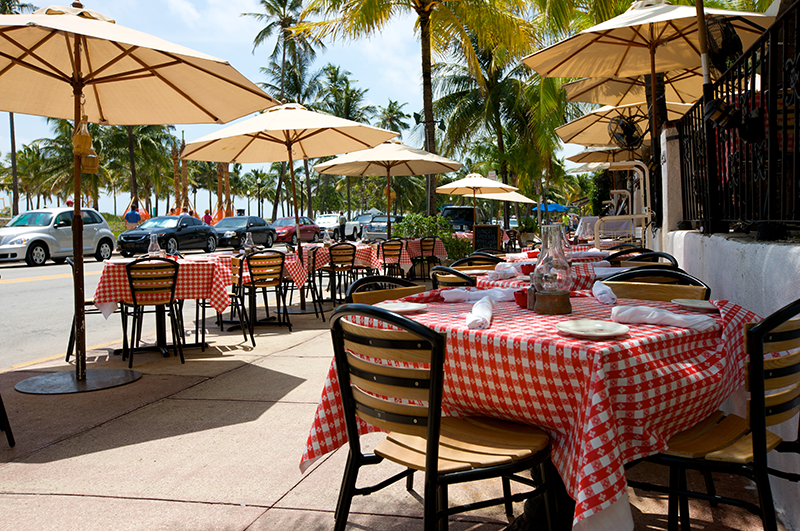 Outdoor restaurant patio with red and white checkered tablecloths, wooden chairs, and umbrellas, located by a street lined with palm trees on a sunny day.