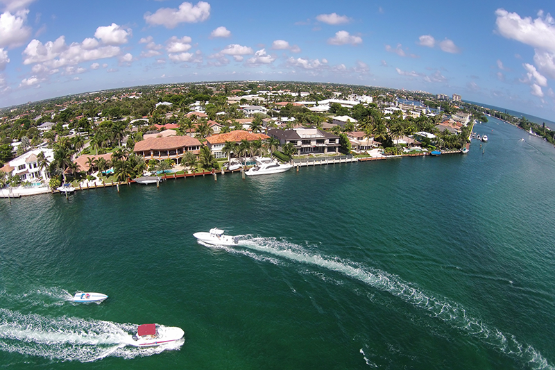 Aerial view of waterfront homes and boats moving along a canal under a partly cloudy sky.