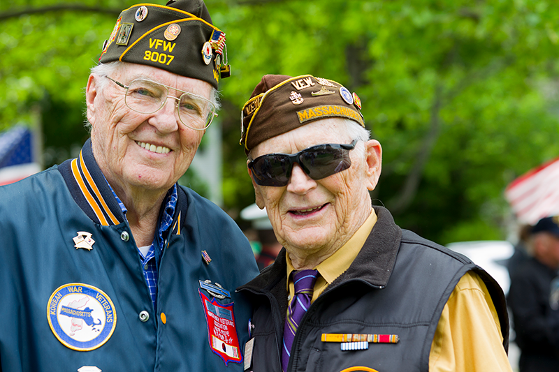 Two elderly veterans wearing military caps and badges stand together outdoors, smiling at the camera with greenery and American flags in the background.