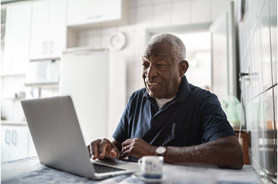 Older man sits at a kitchen table using a laptop, smiling while looking at the screen. The kitchen is bright with white cabinets and a refrigerator in the background.