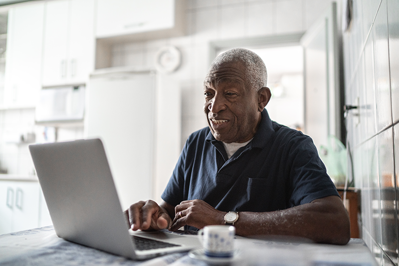 Older man sitting at a kitchen table using a laptop, smiling as he looks at the screen.