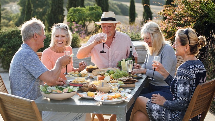 Five adults sit outdoors at a table enjoying food, drinks, and conversation, surrounded by greenery and sunlight.
