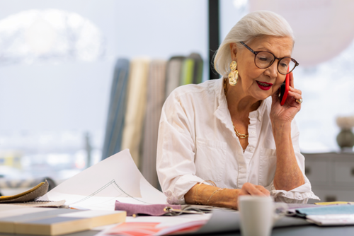 Older woman with gray hair and glasses, wearing a white shirt, talks on a red phone while sitting at a desk covered with papers, fabric samples, and a coffee cup.