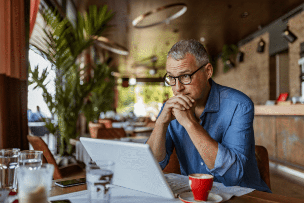Man wearing glasses sits at a table with a laptop and coffee cup, looking thoughtful in a cafe with plants and warm lighting.