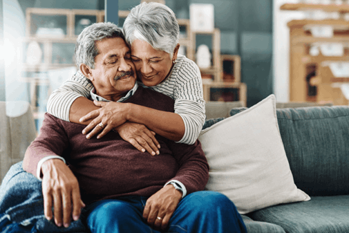An older woman hugs an older man from behind as they sit on a couch in a cozy living room, both appearing content and relaxed.