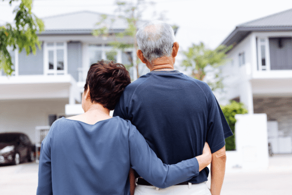 An older couple stands outside, facing away, with arms around each other in front of two modern houses on a residential street.