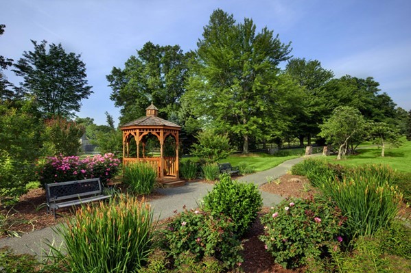 A wooden gazebo stands by a pathway in a landscaped garden with benches, flowering bushes, and tall green trees under a clear blue sky.