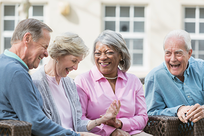 Four older adults sit closely together outdoors, smiling and laughing, with a building and windows visible in the background.