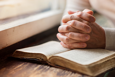 Person with hands clasped in prayer resting on an open book, possibly a Bible, near a window on a wooden surface.