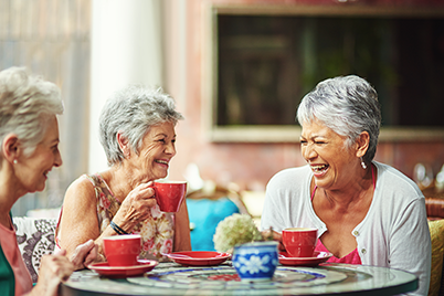 Three older women sit at a round table, smiling and laughing while holding red mugs, with decorative plates and plants in the background.